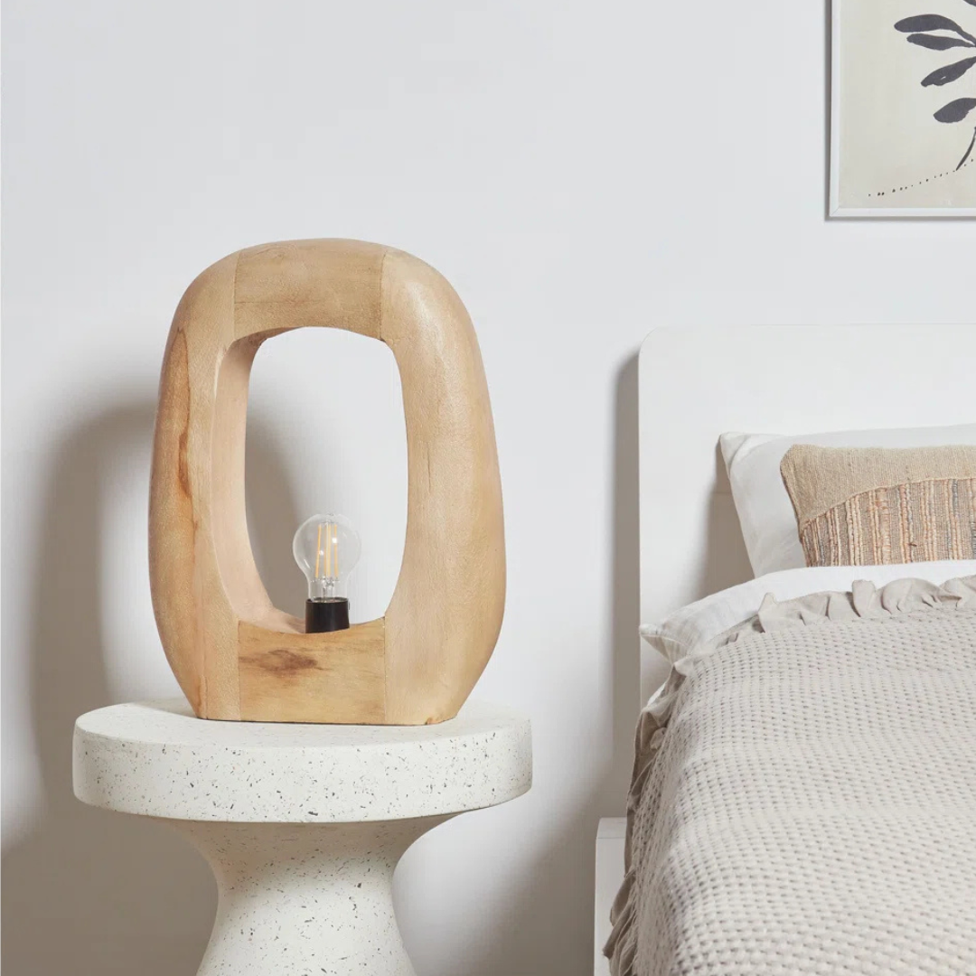 Wooden lamp on a white pedestal next to a bed with a neutral color scheme.