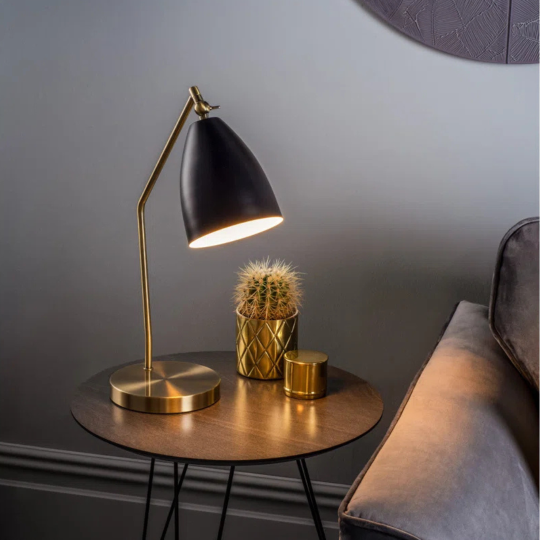 Modern black desk lamp on a small round table with a cactus and candle in a dimly lit room.