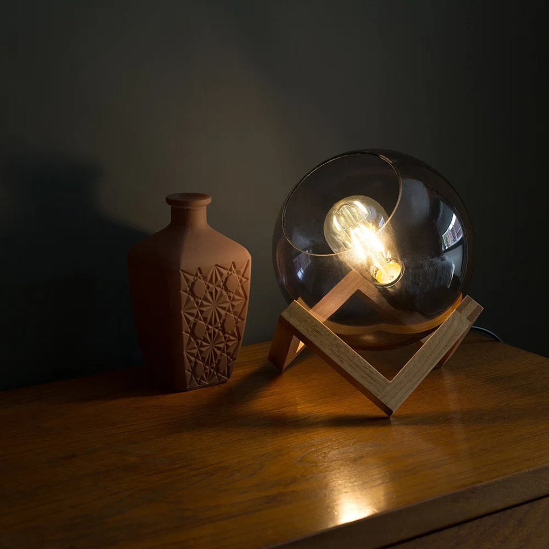 Glass orb with light bulb inside on a wooden stand next to a textured vase on a dark surface