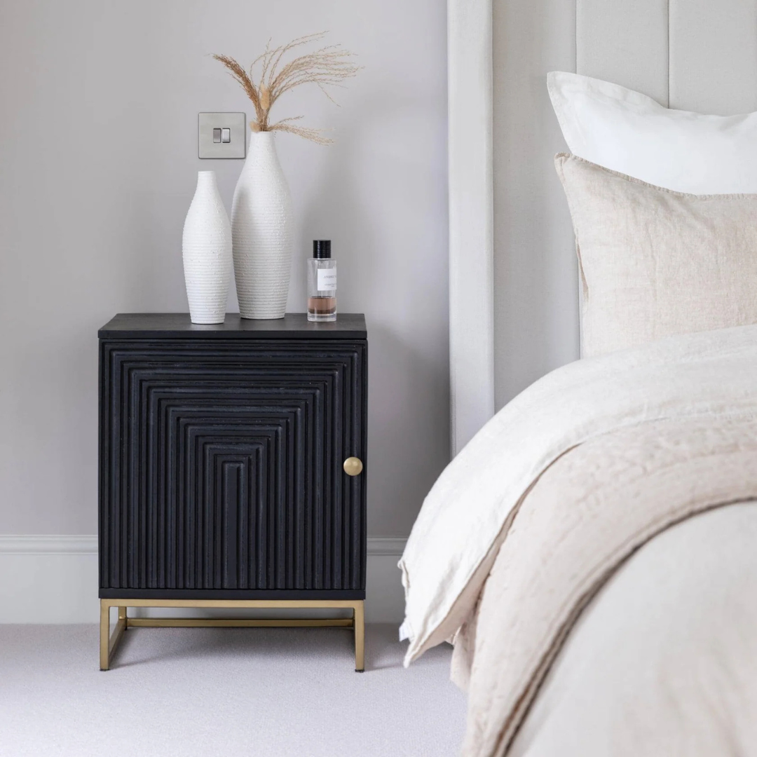 Black textured nightstand with decorative items next to a bed in a bedroom setting.