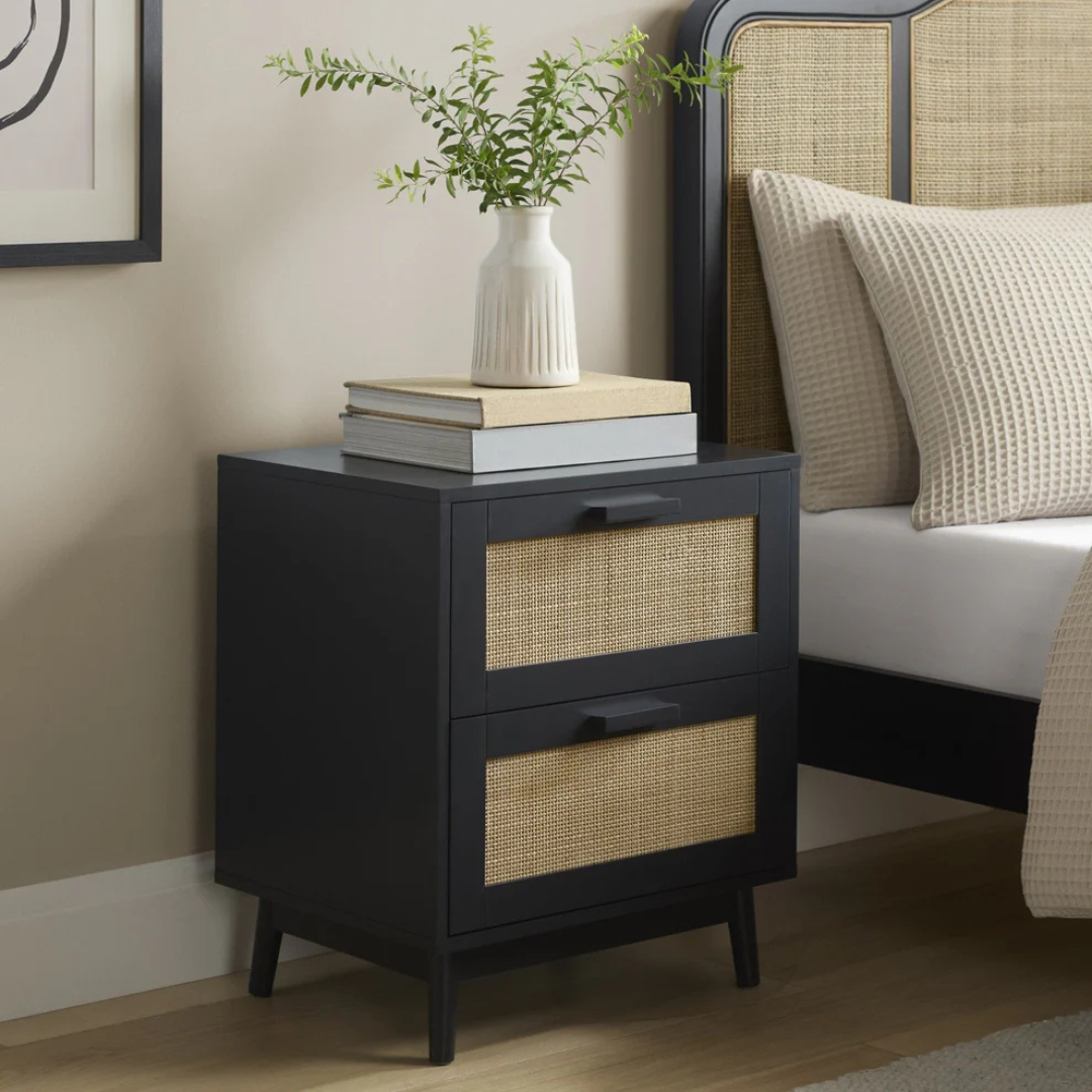 Black nightstand with woven drawers next to a bed with beige pillows.