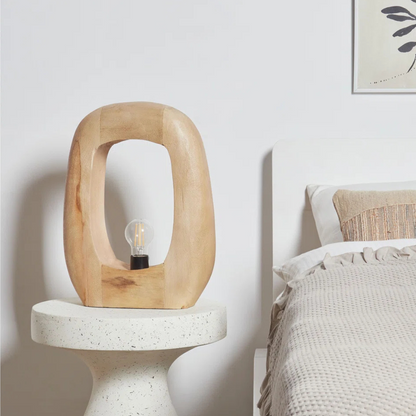 Wooden lamp on a white pedestal next to a bed with a neutral color scheme.