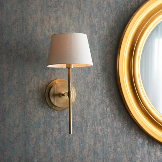 Wall-mounted lamp with a white shade next to a gold-framed mirror on a textured wall.