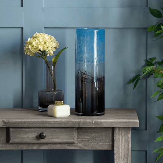 Tall cylindrical vase with gradient design on a wooden table against a blue wall.
