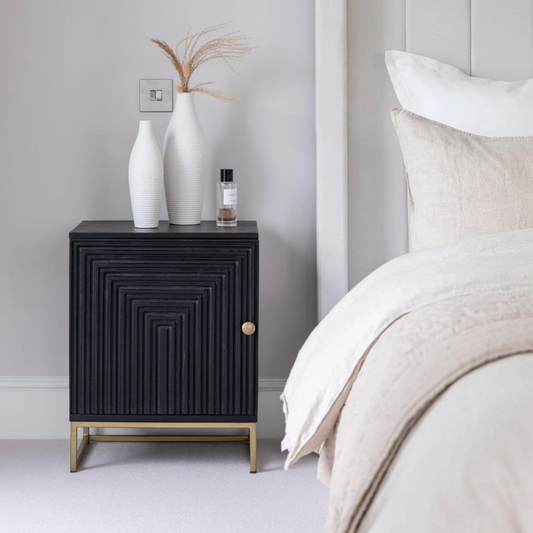Black textured nightstand with decorative items next to a bed in a bedroom setting.