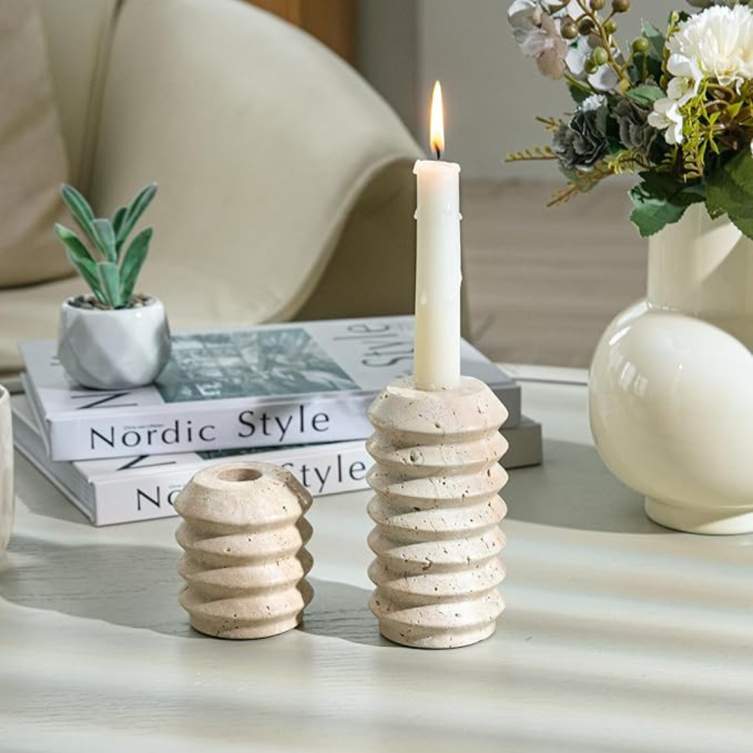 Two ceramic candle holders with a lit candle on a table with books and a plant in the background.
